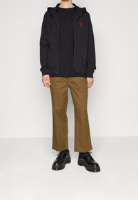 Persona che indossa un hoody nero con zip, una maglietta nera, pantaloni marroni e stivali neri con lacci, in piedi contro uno sfondo chiaro e uniforme.