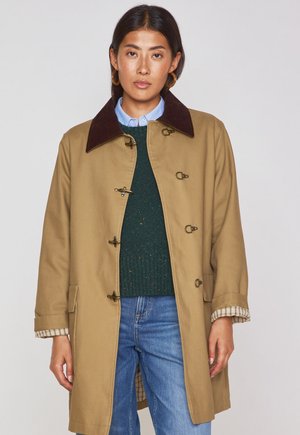 Tan outer coat with a brown corduroy collar, toggle fastenings, and checkered lining, worn over a green sweater and jeans.