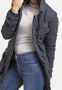 Mörkblå vadderad jacka med hög krage, knäppning fram och sidofickor, bärs över en grå ribbad topp och blåjeans.