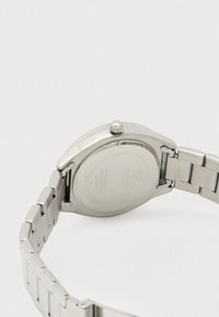 Ure i rustfrit stål med rund kasse, sølvfarvet ledarmbånd og en tekstureret, indgraveret bagside med mærke- og vandafvisningsdetaljer.