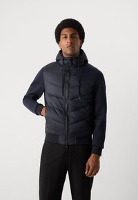 Navy puffer vest with a hood, featuring a zippered front, elastic cuffs, and two side pockets. Textured, quilted upper for insulation.