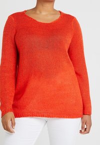Helle orange gestrickte Pullover mit rundem Halsausschnitt, langen Ärmeln und entspannter Passform, kombiniert mit weißen Hosen mit strukturiertem Stich.
