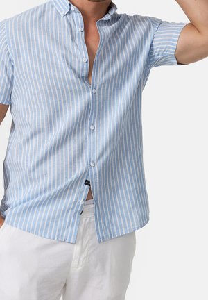 Homme portant une chemise à manches courtes rayée bleu clair et blanc avec un pantalon blanc, posant avec un bras levé.