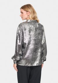 Metallic silver blouse with a textured finish, long sleeves, and a relaxed fit. Features a collar and gathered detailing at the cuffs.