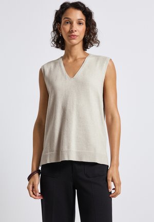 Vrouw met krullend haar, gekleed in een beige mouwloze V-halstrui en een zwarte broek, staand tegen een effen lichte achtergrond.