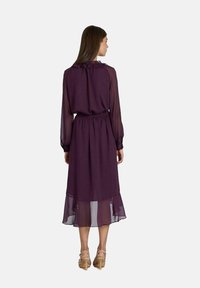 Robe longue et violette en matériau léger, avec un col froncé, une taille élastique et un ourlet superposé. Vue de dos montrée.