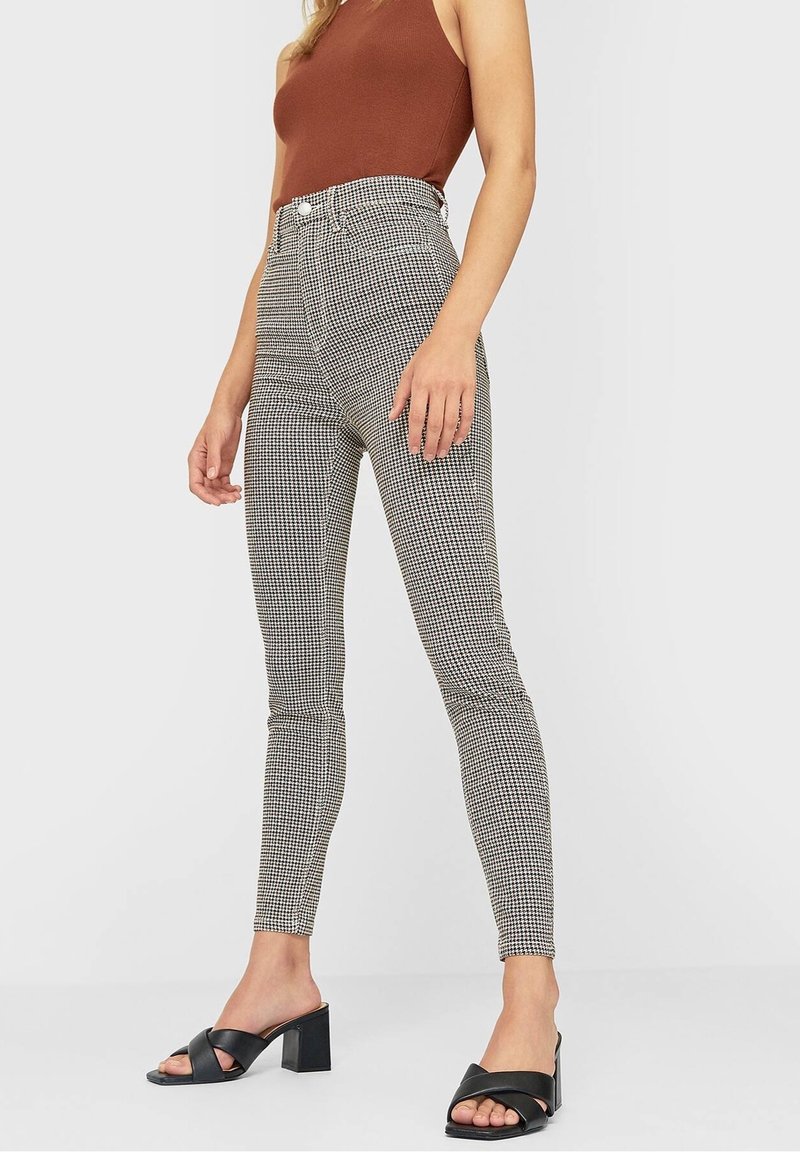 Vrouw in zwart-witte houndstooth slim fit broek met hoge taille, bruin mouwloos topje en zwarte sandalen met hoge hakken en gekruiste bandjes.