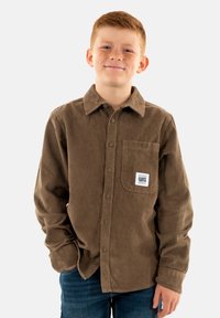 Camisa de pana marrón con cuello abotonado, bolsillo frontal y etiqueta de marca. Textura vertical estriada notable. Llevada con jeans azules.