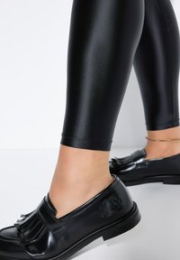 Zwarte glanzende legging gecombineerd met zwarte loafers met franjes. De legging zit strak en heeft een gladde textuur, en reikt tot halverwege de kuit.