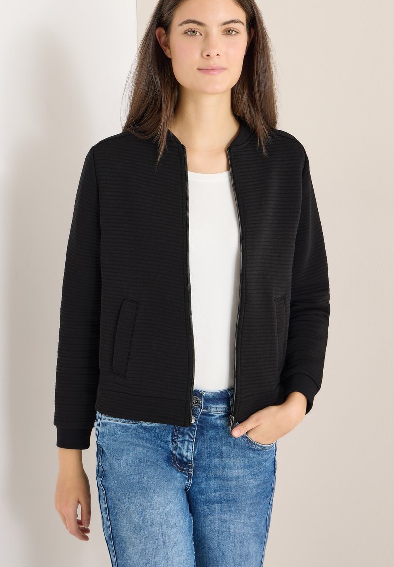 Bomber nero con trama a coste, chiusura con zip e due tasche laterali. Indossato sopra una maglia bianca e abbinato a jeans blu.