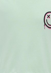 Ανοιχτό πράσινη κοντομάνικη μπλούζα φτιαγμένη από βαμβάκι, με γραφικό σχέδιο ενός μαύρου και ροζ χαμογελαστού προσώπου στο μπροστινό μέρος. Λείος υφασμάτινος υφή.