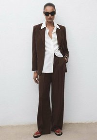 Bruin op maat gemaakt pak met wijde broekspijpen, een witte gestreepte blouse eronder en een paar zwarte zonnebrillen. Rode en zwarte sandalen maken de look compleet.