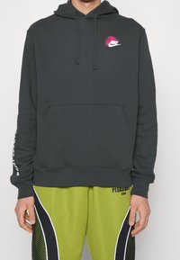 Ciemnoszara bluza z kapturem z przednią kieszenią typu kangurka, regulowanym sznurkiem w kapturze oraz różowym logo Nike na piersi. Materiał wydaje się miękki i teksturowany.