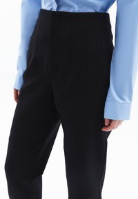 Černé elegantní kalhoty s vysokým pasem, s hladkým povrchem, předními sklady a bočními kapsami, v kombinaci se světle modrou košilí.