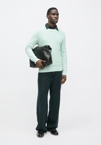 Hellgrüner, gemusterter Strickpullover, dunkelblaue Jeans mit weitem Bein, hält eine schwarze Ledertasche, vor einem schlichten weißen Hintergrund.