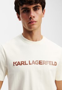 Crème katoenen t-shirt met een prominent weergegeven bruine "KARL LAGERFELD" logo. Klassieke ronde halslijn, korte mouwen, gladde textuur.