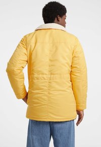 Geel pufferjack met een witte sherpa-kraag, zijzakken en gestikt detail, gecombineerd met lichtblauwe jeans.