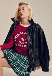 Musta nahkatakki, punainen collegepaita valkoisilla teksteillä ja vihreä ruudullinen hame. Keskeisiä piirteitä ovat materiaalit ja kontrastiset värit.