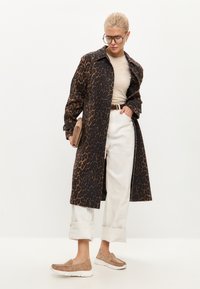 Trenčkot s leopardím vzorem v tmavě hnědé a černé barvě, vrstvený přes béžový top a bílé kalhoty. Hnědé slip-on boty a hnědá kabelka.