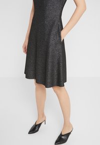 Czarna sukienka bez rękawów z rozkloszowanym dołem, wykonana z błyszczącego materiału. Posiada boczne kieszenie i jest zestawiona z czarnymi, spiczastymi klapkami.