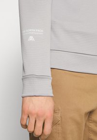 Camisetta grigia chiaro a maniche lunghe con tessuto testurizzato, caratterizzata da una stampa bianca del marchio sul polsino e sull'orlo delle maniche ribattuto.