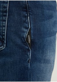 Gros plan sur la poche d'un jean en denim bleu avec une étiquette en tissu noir visible affichant le texte blanc « CHASIN' ».