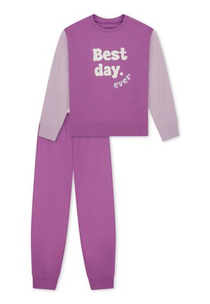 Smiling girl with long blonde hair wearing purple and lavender pajamas with "Best day ever" text, standing barefoot on a plain background.