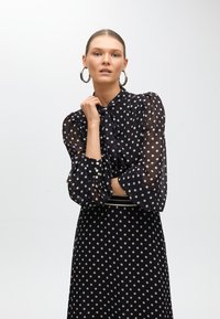 Svart blus med prickar i genomskinligt material, hög krage, knytdetalj och ballongärmar. Matchad med en nederdel i samma prickiga mönster.