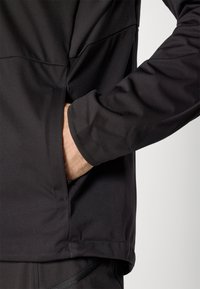 Person's right hand inserted into the side pocket of a black jacket with matching black pants visible below.