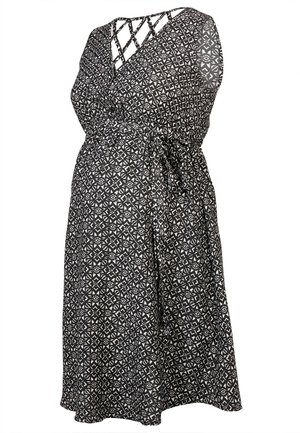 Femme portant une robe sans manches noire et blanche à motifs, longueur genoux, avec un décolleté en V et des chaussures beige à enfiler, debout avec une jambe croisée.