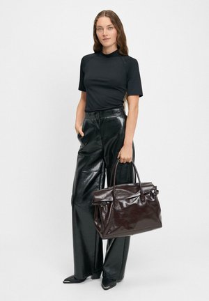 Ciemnobrązowa skórzana torebka o strukturalnym kształcie, z podwójnymi uchwytami i minimalną ilością elementów metalowych. Model nosi czarną bluzkę oraz błyszczące czarne spodnie w kant.