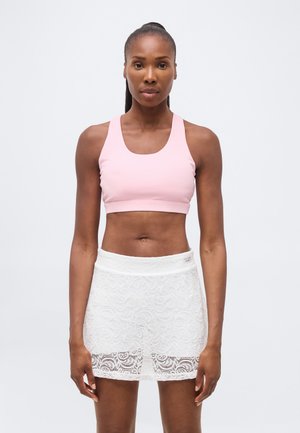 Femeie atletică purtând un sutien sport roz deschis și o fustă albă din dantelă, stând în fața unui fundal simplu și deschis.