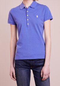 Lila polo-shirt i bomull, med krage, fem knappar i knäppningen och en liten vit logotyp på vänster bröst. Bärs med mörka jeans.
