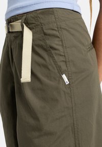 Olive green pants with a textured fabric, featuring a beige belt, side pockets, and a small brand label on the left side.