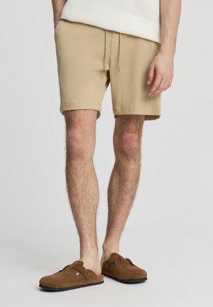 Uomo che indossa pantaloncini beige con coulisse in tessuto strutturato e zoccoli scamosciati marroni senza lacci, in piedi davanti a uno sfondo chiaro e uniforme.