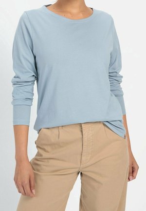 Person trägt hellblaues Langarmhemd, das teilweise in beige Hose gesteckt ist, steht mit entspannten Händen an den Seiten.