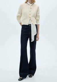 Cárdigan de punto grueso color marfil con botones dorados, combinado con jeans acampanados azul oscuro y zapatos negros, con un lazo blanco en la cintura.