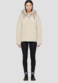 Beżowa kurtka z polaru z błyszącym beżowym kapturem, przednimi kieszeniami i czarnymi sznurkami. Noszona z ciemnymi dżinsami i czarnymi botkami za kostkę.