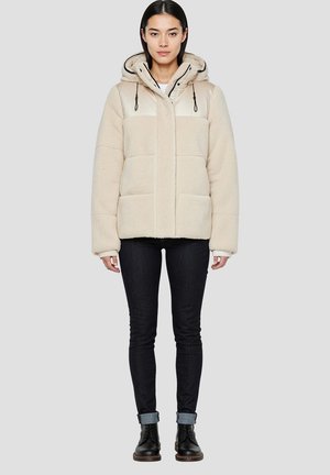 Giacca in pile beige con cappuccio beige lucido, tasche frontali e coulisse nere. Indossata con jeans scuri e stivaletti neri.