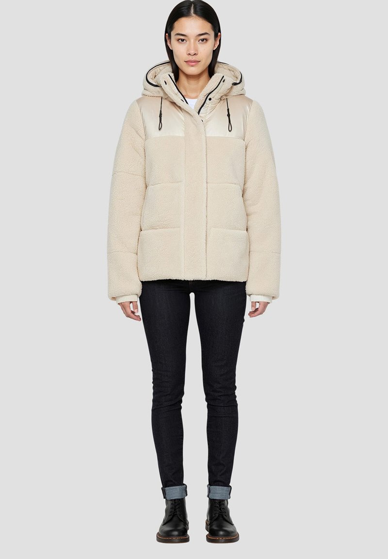 Beżowa kurtka z polaru z błyszącym beżowym kapturem, przednimi kieszeniami i czarnymi sznurkami. Noszona z ciemnymi dżinsami i czarnymi botkami za kostkę.