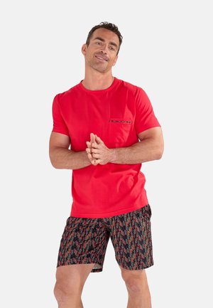 Uomo con T-shirt rossa a maniche corte con taschino sul petto e pantaloncini neri, rossi e bianchi a fantasia, in piedi con le mani intrecciate.