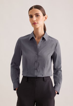 Femme aux cheveux foncés en queue de cheval, portant une chemise grise à boutons et un pantalon noir, debout les mains dans les poches devant un fond uni.