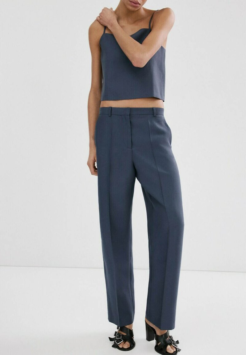 Kvinde iført en ærmeløs mørkegrå crop top, matchende højtsiddende bukser og sorte åbne hælede sandaler med spænder.