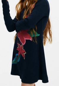 Merensininen ribbineulottu mekko, jossa pitkät hihat ja suuret, värikkäät kukkakuviot vaaleanpunaisina ja vihreinä. Liehuva helma, pehmeä pinta ja rento istuvuus.