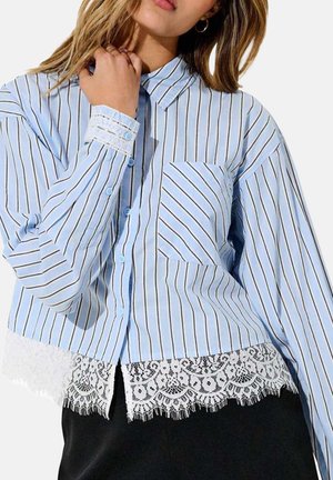 Donna che indossa una camicia a righe azzurro chiaro con bottoni, rifinita con pizzo sui polsini e sull'orlo, abbinata a pantaloni neri.