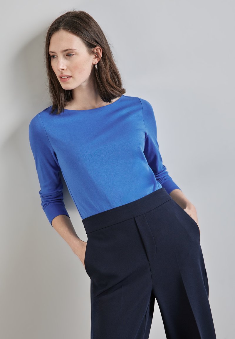 Blå långärmad topp med rund halsringning, ihop med högmidjade marinblå byxor. Slätt tyg med en lätt lyster och skräddarsydd passform.