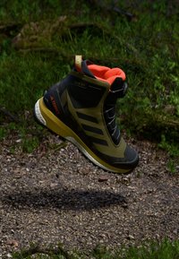 Półwysokie buty trekkingowe w czarnym i oliwkowym kolorze, z jasnopomarańczowym kołnierzem, teksturowaną powierzchnią, warstwową konstrukcją i solidną podeszwą.