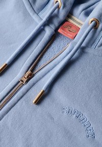 Felpa con zip blu chiaro realizzata in morbido tessuto. Caratterizzata da accessori dorati, cordini e logo ricamato "SUPERDRY". Texture liscia.