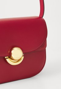 Borsa a tracolla in pelle rossa con pattina arrotondata, chiusura circolare dorata e dettagli in suede a contrasto. Texture liscia con cuciture pulite.