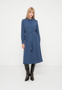 Hosszú ujjú, gombos ruházat mélykék szövetből, derékban megkötővel, gallérral és lágy, középkalás hosszúságú fazonnal. Barna térdig érő csizmák.